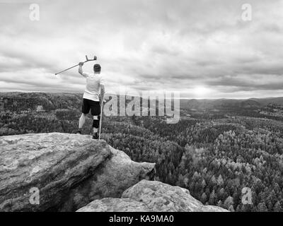 Glückliche Menschen Wanderer holding Medizin Krücke über dem Kopf, verletzte Knie Knie fest programmierte Strebe. Scenic mountain top mit tiefen nebligen Tal unten Stockfoto