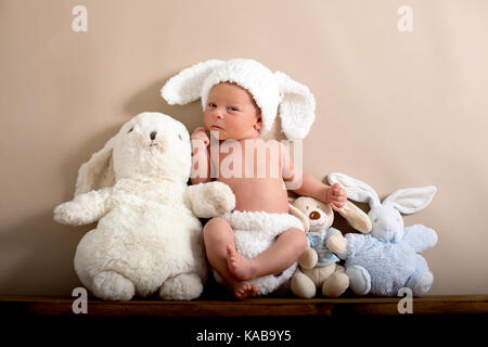 Neugeborenes Baby Boy trug ein braunes Kaninchen hat und Hosen aus Gewirken, schlafen auf einem Regal neben kleinen flauschigen Kaninchen. Im Studio Schuß auf eine cremige Hinterg Stockfoto