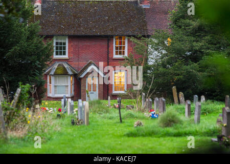 Blick auf dem Friedhof der All Saints Church mit einem roten Backstein Cottage mit, die Beleuchtung im Hintergrund, East Meon, Hampshire, Großbritannien Stockfoto