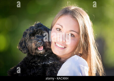 Maltipoo (Maltese x Toy Pudel). Lächelnde Frau Wange an Wange mit Schwarz und tan Hund. Deutschland Stockfoto