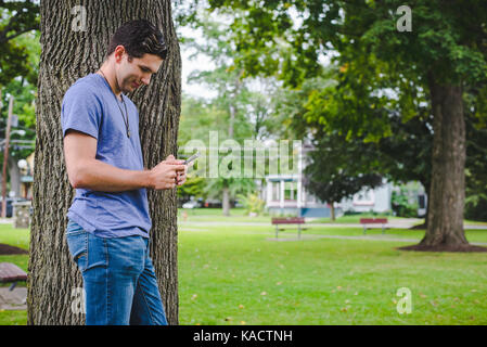 Ein junger Mann schaut auf sein Handy während gegen einen Baum im Park gelehnt. Stockfoto