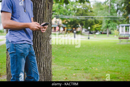 Ein junger Mann schaut auf sein Handy während gegen einen Baum im Park gelehnt. Stockfoto