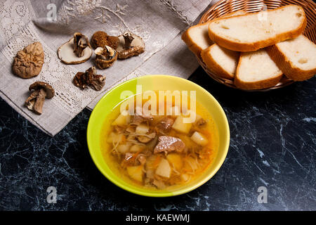 Pilzsuppe in grüne Platte auf einem schwarzen Stein. Mehrere getrocknete Steinpilze oder Weiße Pilze auf braunem Tuch. Weißbrotscheiben in Aalen Stockfoto