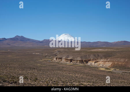 Schnee bedeckt Parinacota Vulkan (6342 m) überragt das Altiplano und Klippen entlang das Tal des Flusses Lauca in den Lauca Nationalpark. Stockfoto