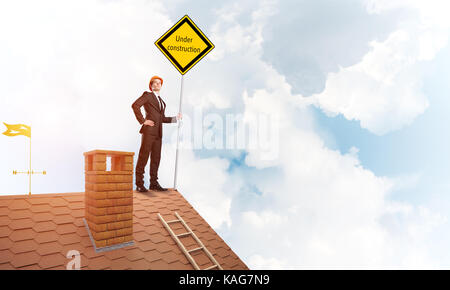 Junge Unternehmer auf Haus Ziegel Dach Holding gelbes Schild. Mixed Media Stockfoto