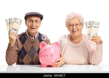 Glückliche Senioren mit Geld-Bundles und ein sparschwein an einem Tisch auf weißem Hintergrund sitzt Stockfoto