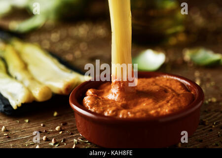 Nahaufnahme von einem gebratenen Calcot, süße Zwiebel typisch für Katalonien, Spanien, eingetaucht in romesco Soße, und einige calcots auf einem rustikalen Holztisch Stockfoto