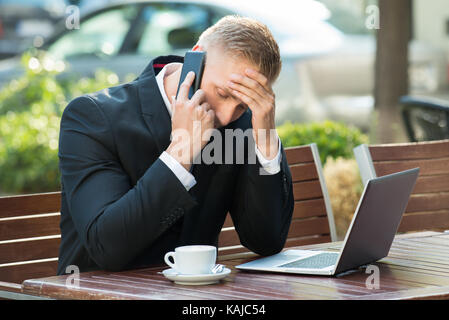 Deprimiert Geschäftsmann Gespräch am Handy mit Kaffeetasse und Laptop Stockfoto