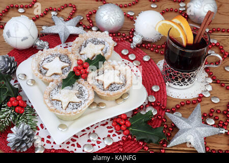 Weihnachten Mince Pies und Stechpalme auf eine herzförmige Platte mit Glühwein und Christbaumkugel Dekorationen auf Eiche Hintergrund. Stockfoto