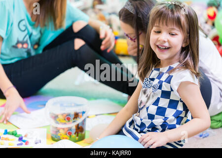 Fröhliche Pre-school Mädchen mit einem trendigen T-Shirt beim Spielen auf dem Fußboden, betreut durch ihre junge und coole Lehrerin in einem modernen Kindergarten Stockfoto