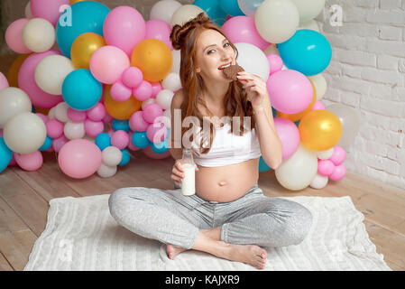 Junge schwangere Frau trinkt Milch sitzen auf dem Boden im Zimmer. Stockfoto