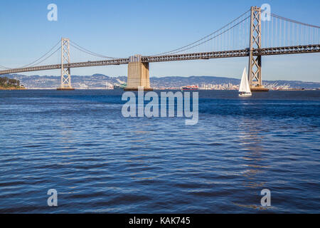 Bay Bridge in San Francisco, Kalifornien, USA. Blaues Wasser im Vordergrund, einzelnes weißes Segelboot über unter Brücke zu passieren. Kopieren Sie Platz im Wasser. Stockfoto