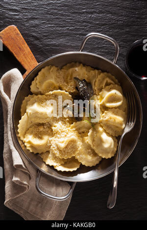 Gekocht serviert pan Ravioli mit Parmesan und gebratenem Salbei. Auf schwarzen Stein Hintergrund mit Glas Rotwein Stockfoto