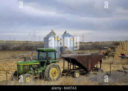 Eine alte John Deere Traktor mit einer rostigen Korn Trailer vor einem Stapel von Heu und zwei Silos. Stockfoto