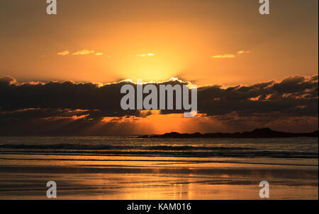 Sonnenaufgang mit Sonne hinter Wolken vor der Küste mit Wasser und Strand im Vordergrund ausgeblendet Stockfoto