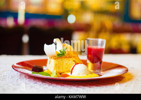 Die rote Platte voller Kuchen, Minze, Ananas mit dem weißen Eis und Saft auf dem Tisch im verschwommenen Hintergrund des Restaurants. Die Nahaufnahme von der Seite. Stockfoto