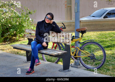 Provinz Fars, Shiraz, Iran - 19 April 2017: Eine modische junge muslimische Frau mit Sonnenbrille und Hijab, lächeln, sitzen auf einer Bank in der Stadt Stockfoto