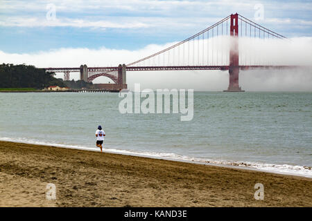 Person Jogging am Strand vor der Golden Gate Bridge, San Francisco, Kalifornien, USA. Brücke ist teilweise im Nebel, Stockfoto