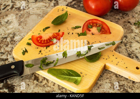 Eine in Scheiben geschnittene Tomaten mit Basilikum auf einem Schneidebrett mit einem kochmesser und ein Bündel von Tomaten auf der Rebe. Stockfoto