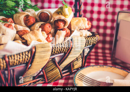 Weidenkorb mit Mini hot dogs Hausgemachte (Würstchen im Teig) und eine Untertasse mit einer Gabel auf einem karierten Hintergrund Stockfoto