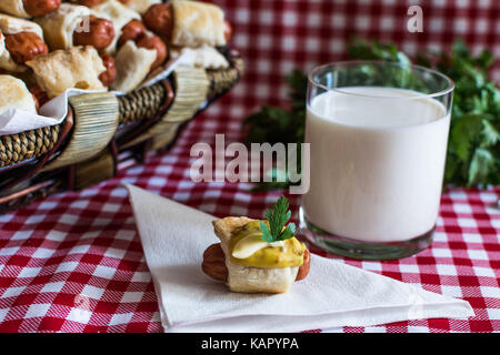 Weidenkorb mit Artisan mini Hot Dogs (Würstchen im Teig) mit Wurst auf eine Serviette in Senf, Mayonnaise und ein Glas frischer Kuh mil abgedeckt Stockfoto