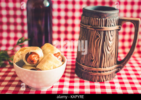 Platte Mini hot dogs Hausgemachte (Würstchen in Blätterteig) auf Serviette mit einer Flasche dunkles Bier und Steingut Tasse auf einem karierten Hintergrund Stockfoto
