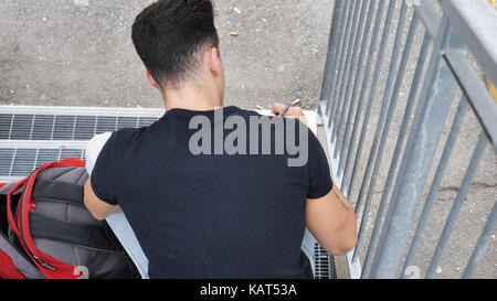 Junger Mann von der Rückseite aus betrachtet, sitzend auf Metall Treppen, das Schreiben von Notizen oder Buchstaben auf Papier mit einem Stift Stockfoto