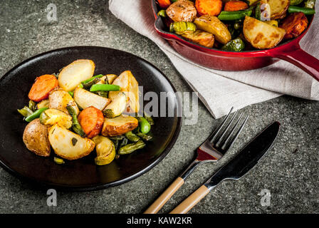 Platte und Pfanne mit gebratenem saisonalen Herbst Gemüse (Zucchini, Kartoffeln, Möhren, Bohnen), auf schwarzen Stein Tabelle kopieren Raum Stockfoto