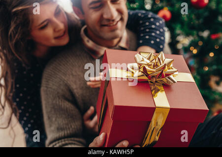Junger Mann und Frau mit einem wunderschön gestalteten Geschenkbox sitzen in der Nähe von einen Weihnachtsbaum. Glückliches Paar Weihnachten feiern mit Geschenken. Stockfoto