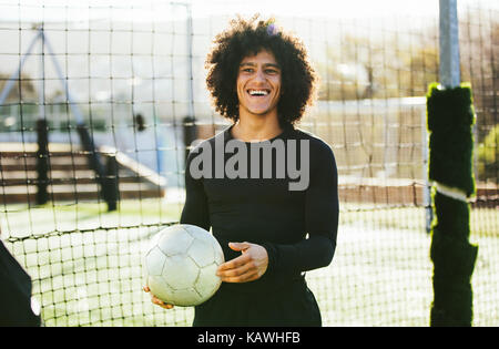 Portrait von Teenage fußball Spieler, der eine Kugel in seiner Hand und lachte. Junger Mann Ausbildung auf dem Fußballfeld. Stockfoto