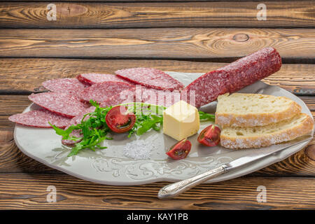 Roh geräucherter Wurst Salami auf weissen Teller, Messer. Zutaten für Sandwiches mit Wurst Thymian, Kräuter, Tomaten, Weißbrot, Butter über Holz- hinterg Stockfoto