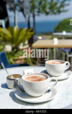 Zwei Cappuccinos auf einem Tisch an der tiefen Hotel Babbacombe Downs, Torquay, Devon, Großbritannien mit einem Fokus auf das Meer im Hintergrund. Stockfoto