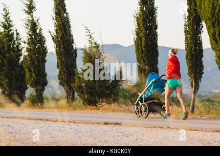 Ausführen von Mutter mit Kind im Kinderwagen bei Mutterschaft bei Sonnenuntergang und Bergwelt. Joggen oder Walken Frau mit Kinderwagen auf bei Sonnenuntergang. Werden Stockfoto