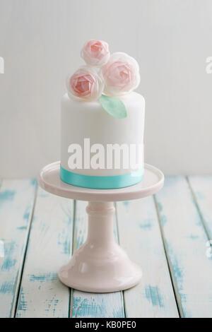 Mini weiß Fondant überzogen Hochzeitstorte mit wafer Papier ranunculus Blumen auf rosa Kuchen stand mit Türkis Ribbon Stockfoto