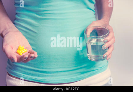 Eine schwangere Frau ersten Trimester in einem blauen T-Shirt hält mehrere Medizin Kapseln, und in der zweiten ein Glas Wasser. IVF Stockfoto