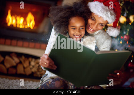 Adorable kleine Mädchen lesen Märchen mit Mama am Heiligen Abend Stockfoto