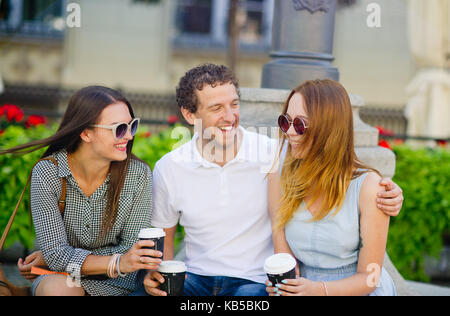 Zwei Mädchen und ein Kerl sind Kaffee trinken und reden lebendig. Freunde auf einer Bank an einem sommerlichen Tag sitzen. Jeder hat eine tolle Stimmung. Stockfoto