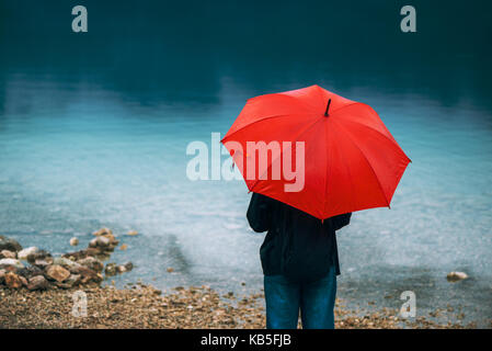 Frau mit roten Dach betrachtet auf Regen vor einem See. Traurig und einsam weibliche Person in die Ferne schauen. Stockfoto