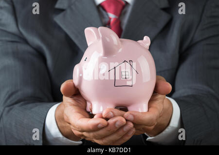 Mittelteil der Geschäftsmann holding Sparschwein mit Haus auf es gezeichnet Stockfoto