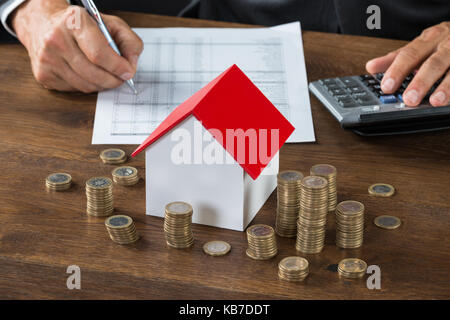 Zugeschnittenes Bild des Geschäftsmannes Berechnung der Steuer von Modell zu Modell Haus und Stapel von Münzen auf Tisch Stockfoto