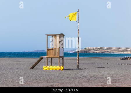 Lonely Rettungsschwimmer Hütte mit gelben Rescue Tube an einem Sandstrand in Teneriffa. Blauer Himmel und Meer im Hintergrund. Eine gelbe Flagge weht über der Hütte Stockfoto