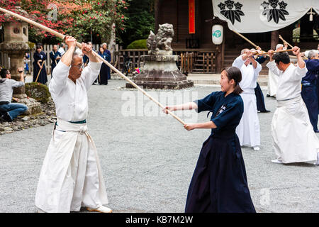 Japan, Osaka, Tada. Master samurai Schwertkämpfer in Weiß im Kampf mit Frau in Schwarz gekleidet, mit bokken oder bokuto, Holz- Schwerter im Heiligtum. Stockfoto