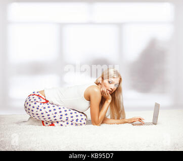 Junge blonde Frau im Schlafanzug auf weißen ganz-Teppichboden Surfen Laptop in der Nähe der Fenster sitzen Stockfoto