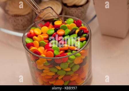 Grün, Braun, Rot und Gelb gefärbte Bonbons in ein Glas mit Cupcakes im Hintergrund Stockfoto