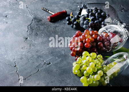 Vielzahl von drei Art frische reife Trauben dunkel Blau, Rot und Grün in verschiedenen Lügen Weingläser mit alten Korkenzieher über schwarzen Textur Hintergrund mit Stockfoto