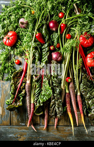 Raw organic lila Karotte mit Vielfalt von Gemüse, Tomaten, Pfeffer, Mangold, Knoblauch, Zwiebel auf Haulm über alte Holzbrett Hintergrund. Ansicht von oben. Essen Stockfoto