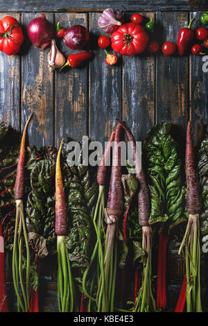 Raw organic lila Karotte mit Mangold Blätter und die Vielfalt an Gemüse Tomaten, Paprika, Knoblauch, Zwiebel über alte Holzbrett Hintergrund. Ansicht von oben mit der Stockfoto