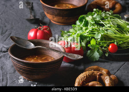 Ton Schüsseln mit vegetarischen hot Möhre tomate Erbse Kartoffel Suppe mit frischem Koriander serviert, Brezeln Brot und Tomaten auf textile Serviette über alte Holz Stockfoto