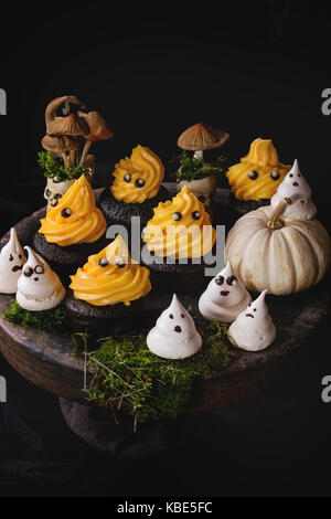 Halloween süßes Dessert Tabelle schwarz Cupcake mit orange Creme, weiß Baiser Geister mit Schokolade Augen, Dekor Schädel und Kürbis auf Lehm p Stockfoto