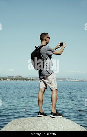 Nahaufnahme eines jungen kaukasischen Mann, der einen Rucksack nehmen Sie ein Foto auf das Meer an der Costa Smeralda, Sardinien, Italien, mit seinem Smartphone Stockfoto